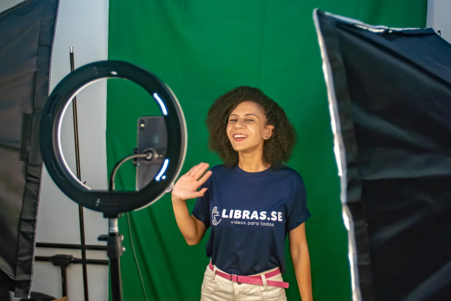 Intérprete de Libras sorrindo e sinalizando em estúdio com camiseta da LIBRAS.SE